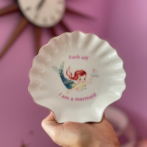 May include: White seashell-shaped trinket dish with a gold rim. The dish features a cartoon mermaid illustration and the phrases "Fuck off" and "I am a mermaid" in pink text. The dish is held in a hand.