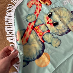 Pode incluir: Uma saia redonda para &aacute;rvore de Natal azul claro com franja de croch&ecirc; branca. Apresenta uma ilustra&ccedil;&atilde;o vintage de um urso azul segurando uma bengala doce, uma rena e um floco de neve.