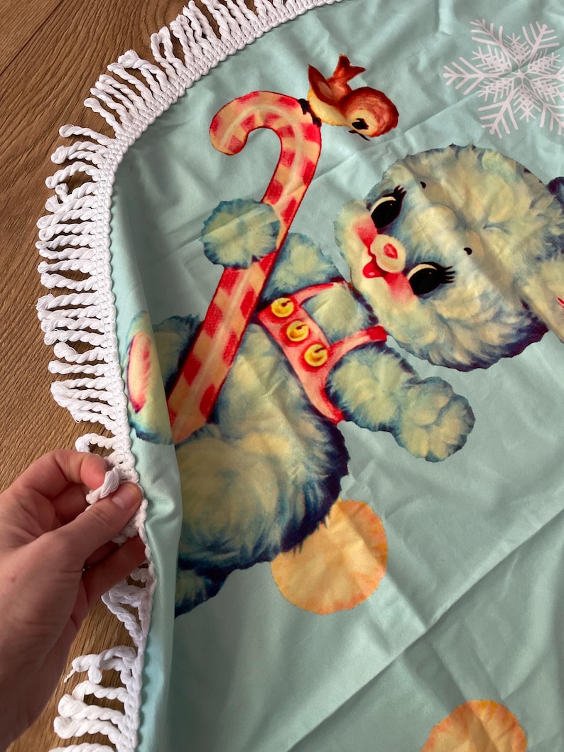 Pode incluir: Uma saia redonda para &aacute;rvore de Natal azul claro com franja de croch&ecirc; branca. Apresenta uma ilustra&ccedil;&atilde;o vintage de um urso azul segurando uma bengala doce, uma rena e um floco de neve.
