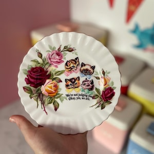 May include: A white scalloped ceramic plate featuring a floral arrangement of red, pink, and yellow roses. Four kittens with colourful bows are illustrated in the centre, accompanied by the printed text: We're not judging you. Okay, yes, we are.