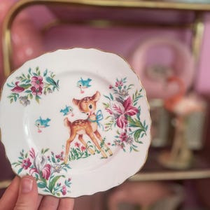 May include: A small, white decorative plate with a gold rim. The plate features a fawn with a blue bow, blue birds, and floral designs in pink, purple, and green. The plate is held in a person's hand.
