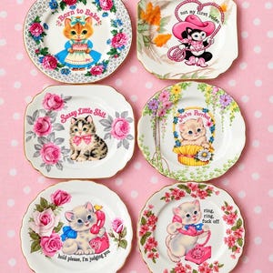 May include: Six white ceramic plates with gold rims, each featuring a kitten illustration and text: Born to Bake, not my first rodeo, Sassy Little Shit, You're Purrfect, hold please, I'm judging you, and ring, ring, fuck off, surrounded by floral motifs.