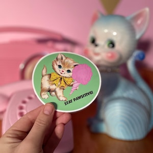 Pode incluir: Um autocolante redondo com uma ilustração vintage de um gatinho segurando algodão doce rosa e o texto "STAY PAWSITIVE!". O autocolante está em frente a uma estatueta de gato azul claro e um fundo rosa.