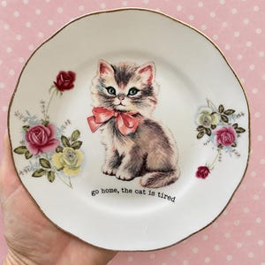 May include: A white ceramic plate with a gold-trimmed scalloped edge, featuring a central illustration of a fluffy grey kitten wearing a pink bow. The plate is decorated with pink and yellow roses and the text, go home, the cat is tired.