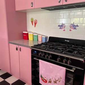 以下が含まれることがあります： ピンク色のキッチン。黒いオーブンとピンク色のクリスマスをテーマにしたタオルがあります。キッチンにはピンク色のキャビネットと白黒の市松模様の床があります。カウンターにはカラフルな容器があり、白いタイルのバックにはアイスクリームコーンと魚のステッカーが貼られています。