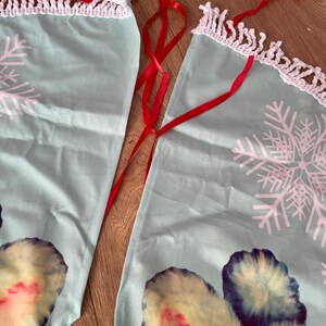 Pode incluir: Duas saias de &aacute;rvore de Natal de tecido azul claro com desenhos de flocos de neve brancos e uma borda de croch&ecirc; branca. Fitas vermelhas s&atilde;o anexadas para amarrar. A parte inferior das saias apresenta um design colorido.