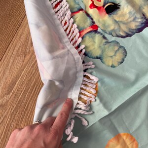 Pode incluir: Uma toalha de praia azul clara com uma ilustra&ccedil;&atilde;o em estilo vintage de um ursinho com olhos pretos e detalhes vermelhos. A toalha tem uma borda de franjas brancas e um fundo azul claro. A toalha &eacute; feita de um material macio.