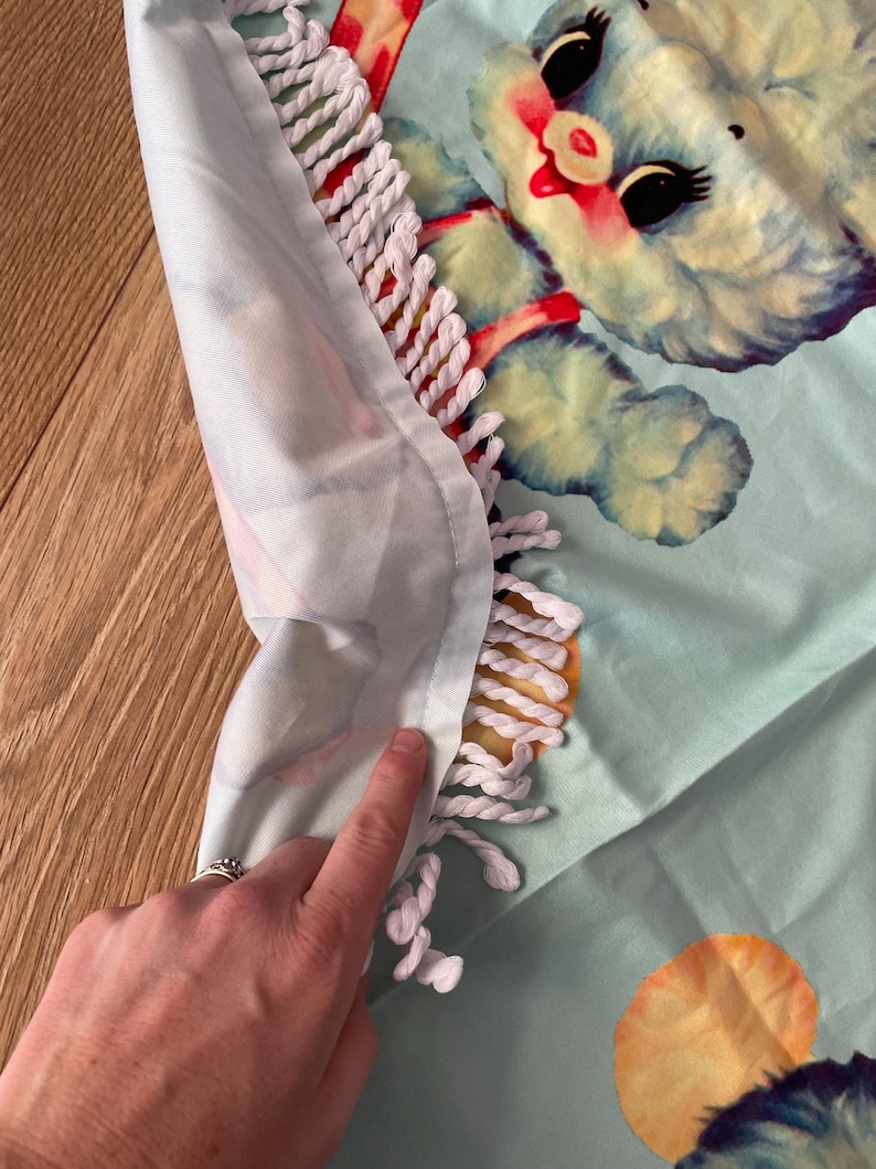 Pode incluir: Uma toalha de praia azul clara com uma ilustra&ccedil;&atilde;o em estilo vintage de um ursinho com olhos pretos e detalhes vermelhos. A toalha tem uma borda de franjas brancas e um fundo azul claro. A toalha &eacute; feita de um material macio.