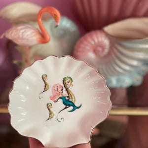 May include: A white, scalloped-edge dish with a gold trim, featuring a mermaid and seahorse design in pink, green, and blue. The dish is held in a hand, with a pink background and decorative items in the background.
