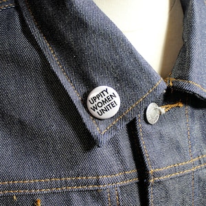 Può includere: Un bottone bianco con il testo "UPPITY WOMEN UNITE!" appuntato su una giacca di jeans blu.
