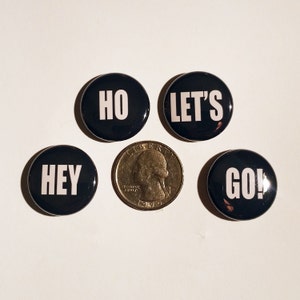 May include: Four black button pins with white text. The pins read "Hey", "Ho", "Let's", and "Go!"