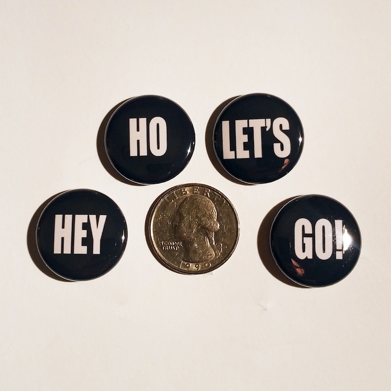 May include: Four black button pins with white text. The pins read "Hey", "Ho", "Let's", and "Go!"