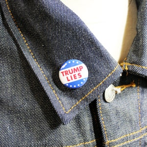 Può includere: Un bottone blu e rosso con stelle bianche attorno al bordo. Il bottone dice "TRUMP LIES" in lettere bianche.