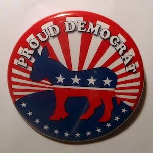 May include: A red, white and blue button with the text "Proud Democrat" and a silhouette of a donkey with three white stars on its side.