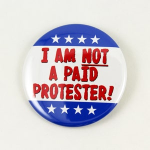 May include: A blue and white button with red text that reads "I AM NOT A PAID PROTESTER!" The button has five white stars on the top and bottom.