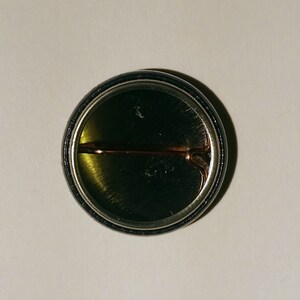 May include: The back of a round button with a black and silver border. A metal pin is attached to the back of the button.