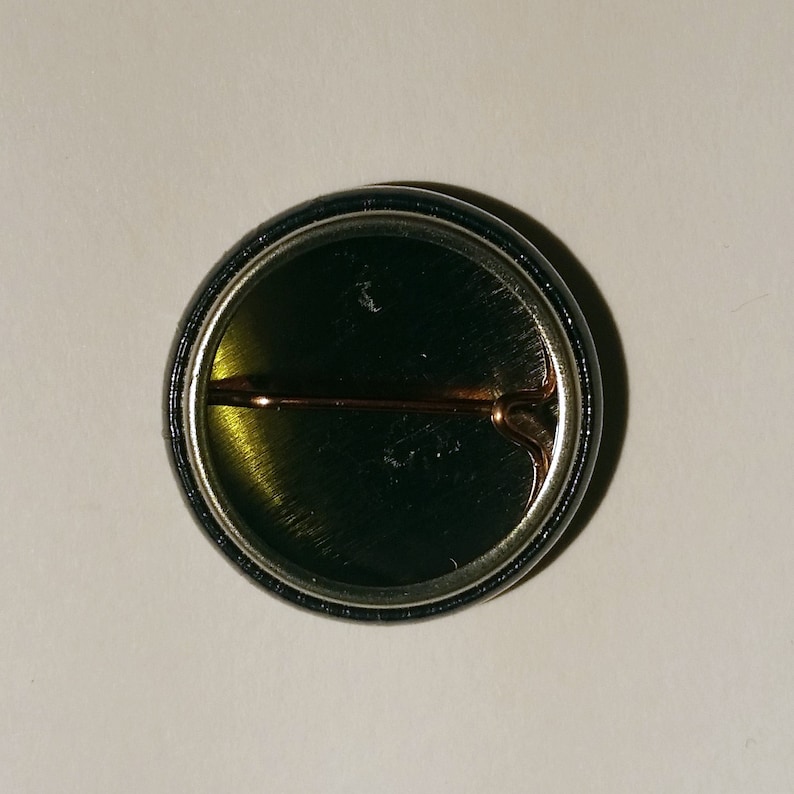 May include: The back of a round button with a black and silver border. A metal pin is attached to the back of the button.