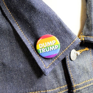 May include: A rainbow-colored button with the text "DUMP TRUMP" in white letters. The button is pinned to a denim jacket.