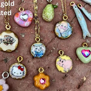 Könnte beinhalten: Eine Sammlung von 14k vergoldeten Anhängern an Goldketten. Die Anhänger zeigen verschiedene Designs, darunter ein Fisch, eine Katze, ein Baby, ein Schwein, ein Herz und eine Blume. Die Anhänger sind farbenfroh und detailliert.