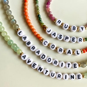 May include: Five beaded bracelets with gemstone beads and white letter beads spelling out the names "Aventurine", "Labradorite", "Coral", "Jasper", and "Ruby".