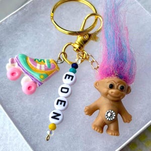 Könnte beinhalten: Ein goldfarbener Schlüsselanhänger mit einem Rollschuh-Anhänger, einer Perlenkette mit der Aufschrift "EDEN" und einer Troll-Puppe mit rosa und blauem Haar. Der Rollschuh ist pastellfarben mit rosa Rollen.