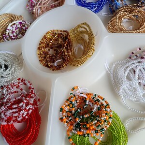 May include: A selection of colourful beaded strands, including gold, brown, red, white, green, orange, and blue, are arranged in a white divided tray. The beads appear to be small and strung together, suggesting they are for jewellery or craft projects.