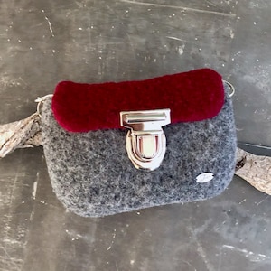 Könnte beinhalten: Eine kleine graue Filmtasche mit einer bordeauxroten Filzklappe und einem silbernen Verschluss. Die Tasche hat einen Riemen und ein kleines Etikett auf der Vorderseite.