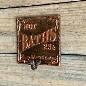 May include: A vintage-style metal sign with a copper finish, advertising "HOT BATHS 25c" with the words "Soap & Towel extra" below. The sign has a decorative border and a hook on the bottom.