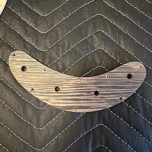 Puede incluir: Un golpeador de guitarra de madera marrón con un patrón de veta de madera negra y varios agujeros para tornillos.