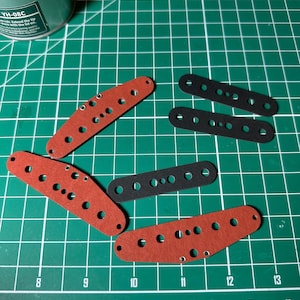 May include: A top-down view of red and black rectangular components with multiple holes, likely for a craft project. They are laid out on a green cutting mat with grid lines and numbers. A small container of solder is visible in the top left corner.