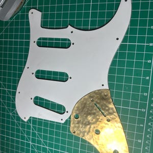May include: A white guitar pickguard with three pickup slots and mounting holes, alongside a textured brass control plate. The pickguard has a classic shape, and the brass plate adds a unique, vintage aesthetic. The items are on a green cutting mat.