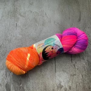 May include: A skein of hand-dyed yarn in vibrant colors. The yarn transitions from orange to pink and purple. A label with text and a colorful design is wrapped around the center of the yarn.