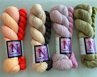 May include: Four sets of yarn skeins, each featuring a main color paired with a smaller accent skein. Colors include peach and orange, cream and dark brown, light pink and rose, and olive green. Each set has a label with the letter N and Nanette Wake Studio.