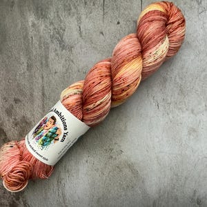 May include: A skein of hand-dyed yarn in shades of peach, orange, and yellow. The yarn is twisted and has a speckled appearance. A white label with the text "Ambitions Yarn" and a colorful illustration is wrapped around the yarn.