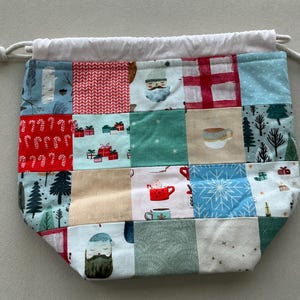 May include: A drawstring pouch featuring a patchwork design of various holiday-themed fabrics. The squares display patterns of candy canes, wrapped gifts, pine trees, mugs of cocoa, snowflakes, and Santa Claus faces in red, green, blue, and white.