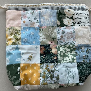 May include: A drawstring pouch featuring a patchwork design of various fabric squares. The patterns include floral prints, woodland animals like rabbits and squirrels, botanical illustrations, and cursive script text in shades of green, blue, yellow, and white.