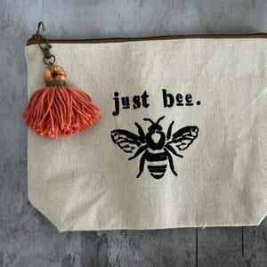 May include: A beige canvas zippered pouch with a black bee graphic and the words "just bee." An orange tassel hangs from the zipper. The pouch is rectangular with a brown zipper and trim.