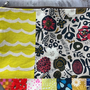 May include: A rectangular fabric pouch with a zipper closure. The pouch is divided into sections with different patterns. One section has a yellow wavy pattern, and another features a floral design with red, navy, and white flowers. The bottom has a patchwork design.