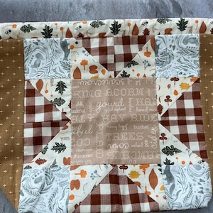 May include: A drawstring bag with a patchwork design. The bag features a brown and white gingham pattern, along with panels of fabric with fall-themed illustrations and text. The bag's exterior is a light brown with a cross pattern.