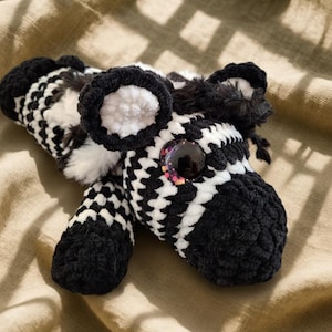 May include: A handmade zebra plush toy with a black and white checkered pattern. The zebra has large, round eyes with colorful accents, fluffy ears, and a black mane. The plush is made of soft, textured yarn.