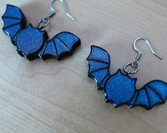 May include: A pair of bat-shaped earrings. Each earring features a dark blue bat with outstretched wings and a black outline. The earrings have silver-colored fish hook ear wires.