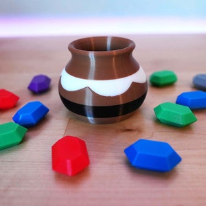 May include: A brown and white ceramic pot with a black band around the middle, surrounded by colourful gemstones. The gemstones are red, blue, green, purple, and grey.