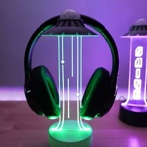May include: A black headset with green LED lights sits on a UFO-shaped stand. The stand has a white base and a green light that illuminates the headset. A second UFO-shaped stand with a purple light is in the background.