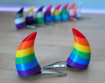 May include: Two rainbow-colored devil horn hair clips with silver metal clasps. The clips are on a light brown wooden surface.