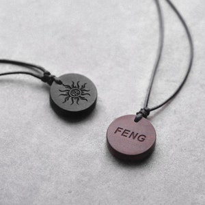 May include: Two wooden pendants on black cords. One pendant is black and features a sun design. The other pendant is brown and has the word "FENG" engraved on it.
