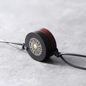 May include: A black and brown wooden pendant with a sun design on the front. The pendant is attached to a black cord.