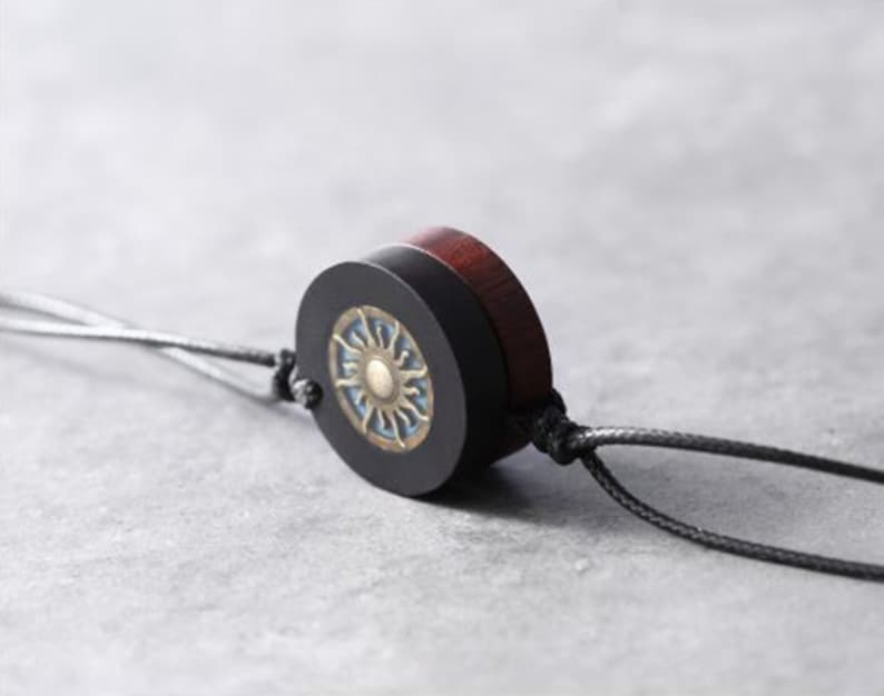 May include: A black and brown wooden pendant with a sun design on the front. The pendant is attached to a black cord.