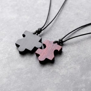 May include: Two black cord necklaces with wooden puzzle piece pendants. One pendant is black and the other is a reddish brown color.