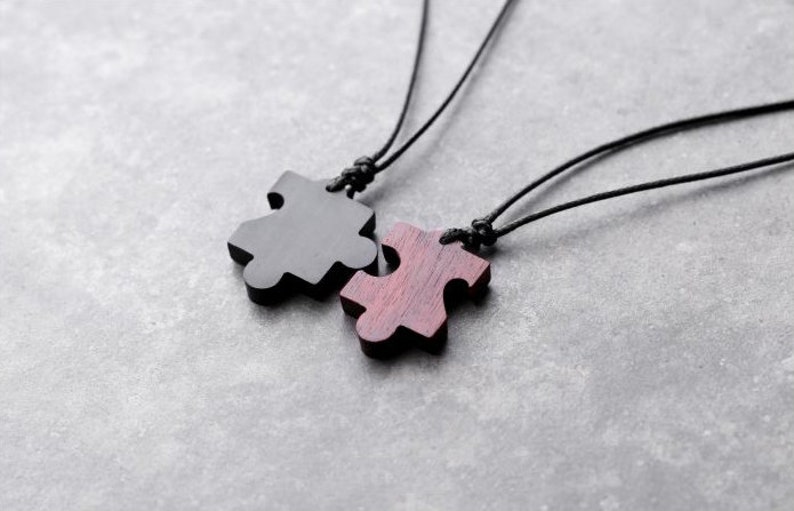 May include: Two black cord necklaces with wooden puzzle piece pendants. One pendant is black and the other is a reddish brown color.