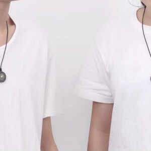 May include: Two people wearing white t-shirts with black necklaces and round pendants. The pendants have a dark wood finish with a gold design.